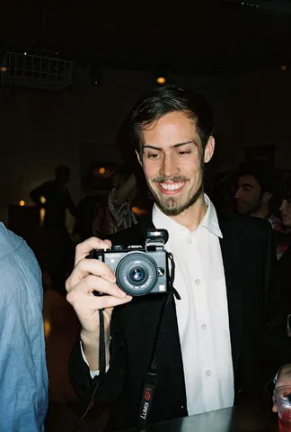Smiling at a dim party in a white shirt and dark blazer, holding a Lumix camera