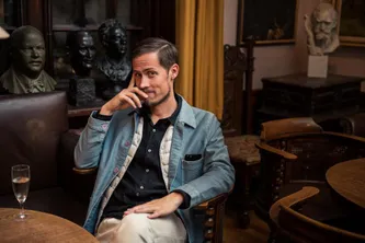 In a denim jacket, phone to ear, seated in a dark wood-panelled room with bronze busts