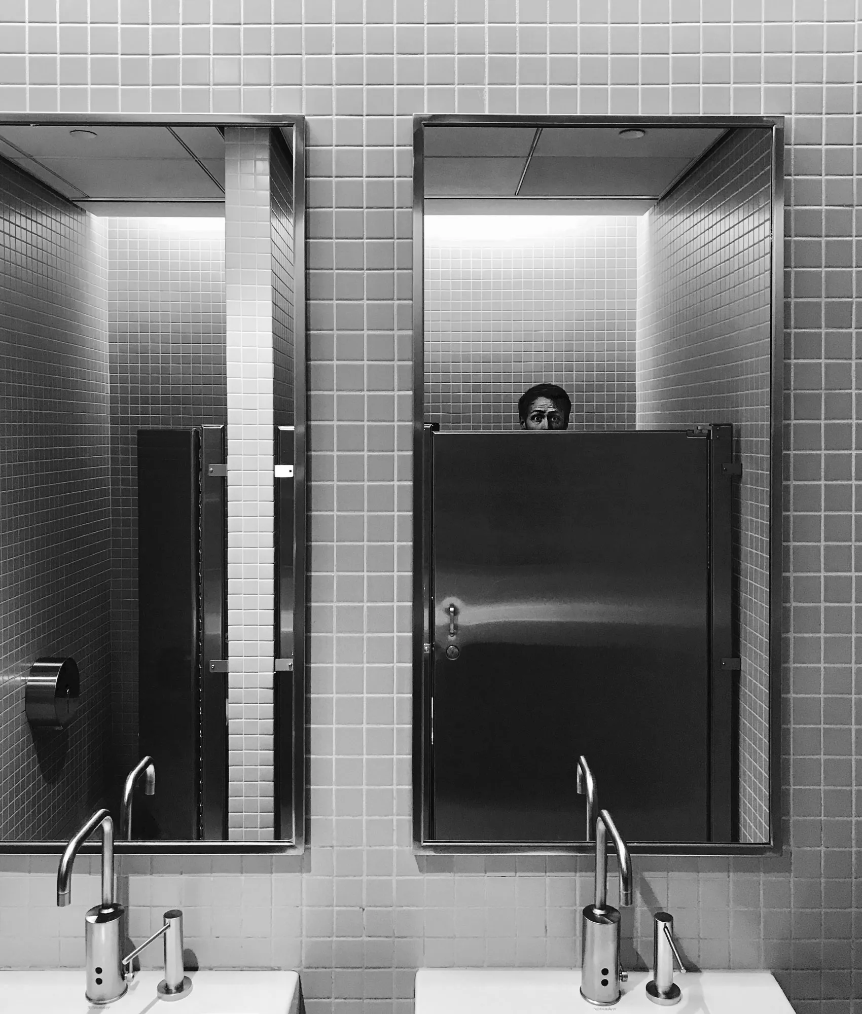 Black-and-white restroom mirrors; in one, Edvard peeks over a stall door