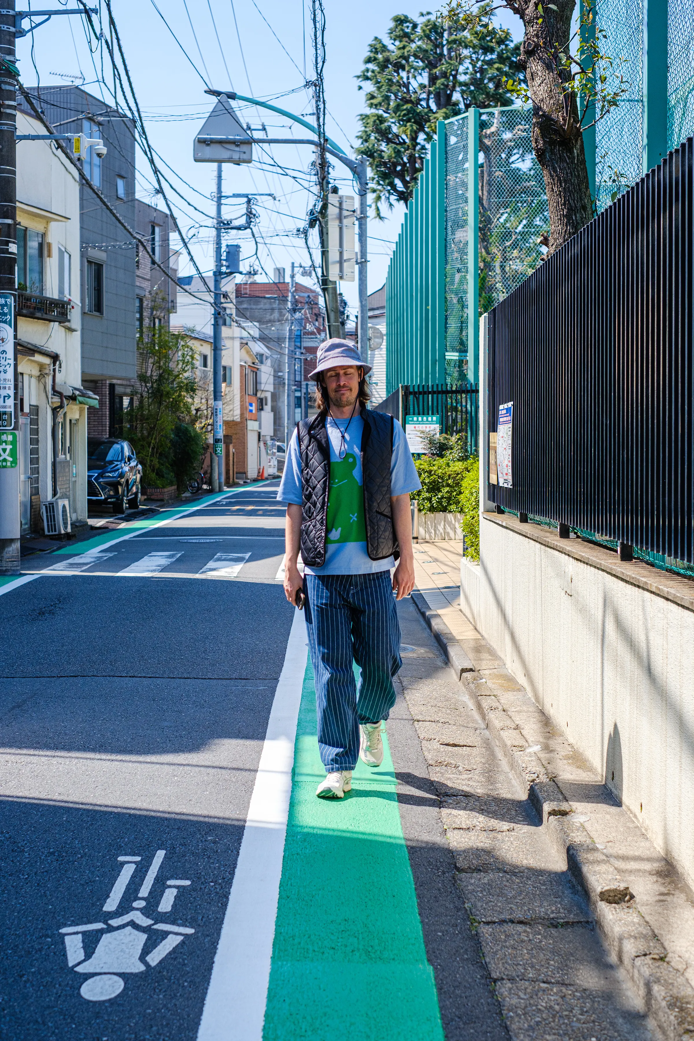 Edvard in a bucket hat, quilted vest and striped trousers, walking a green-striped lane down a sunny side street