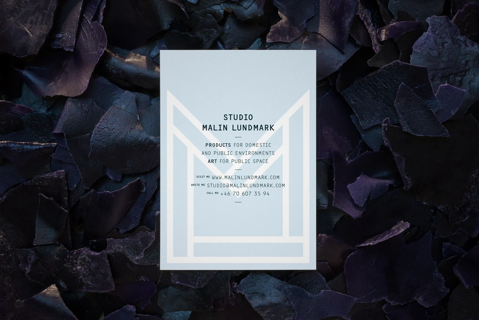 Pale blue card with a large outlined M behind the studio details, on dark purple flakes