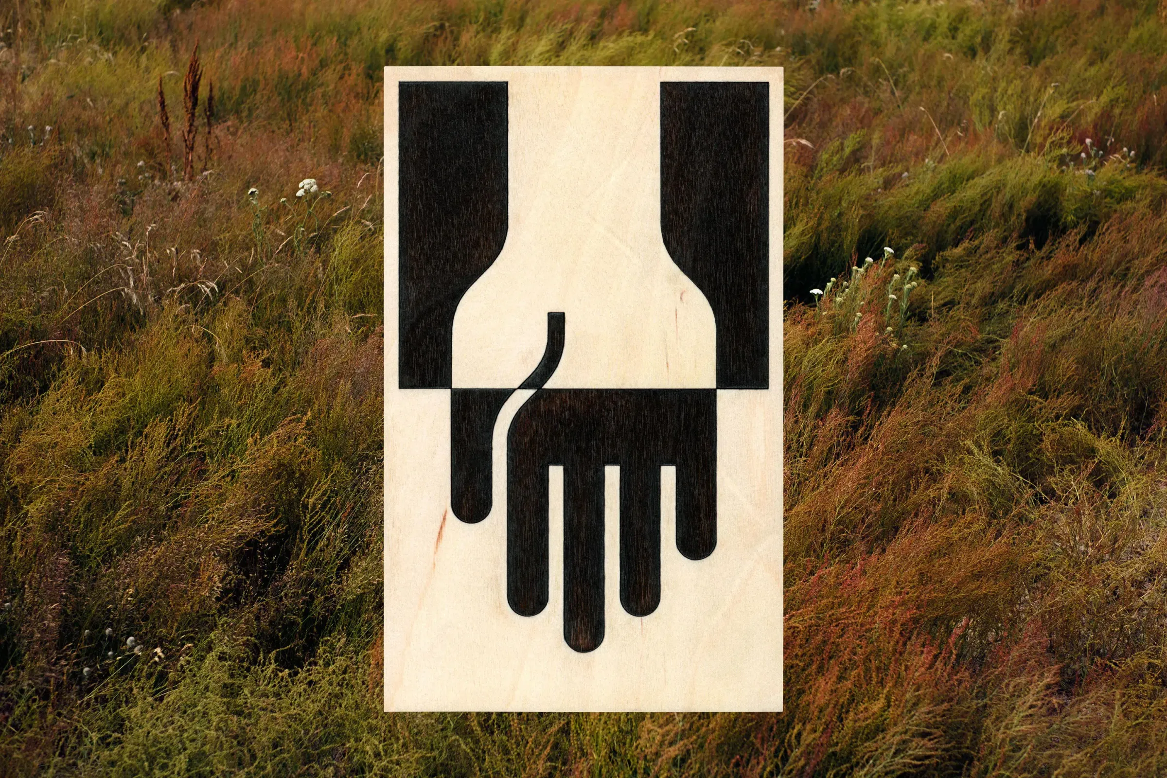 Pictogram of one hand holding another, burned into plywood, set against windswept meadow grass
