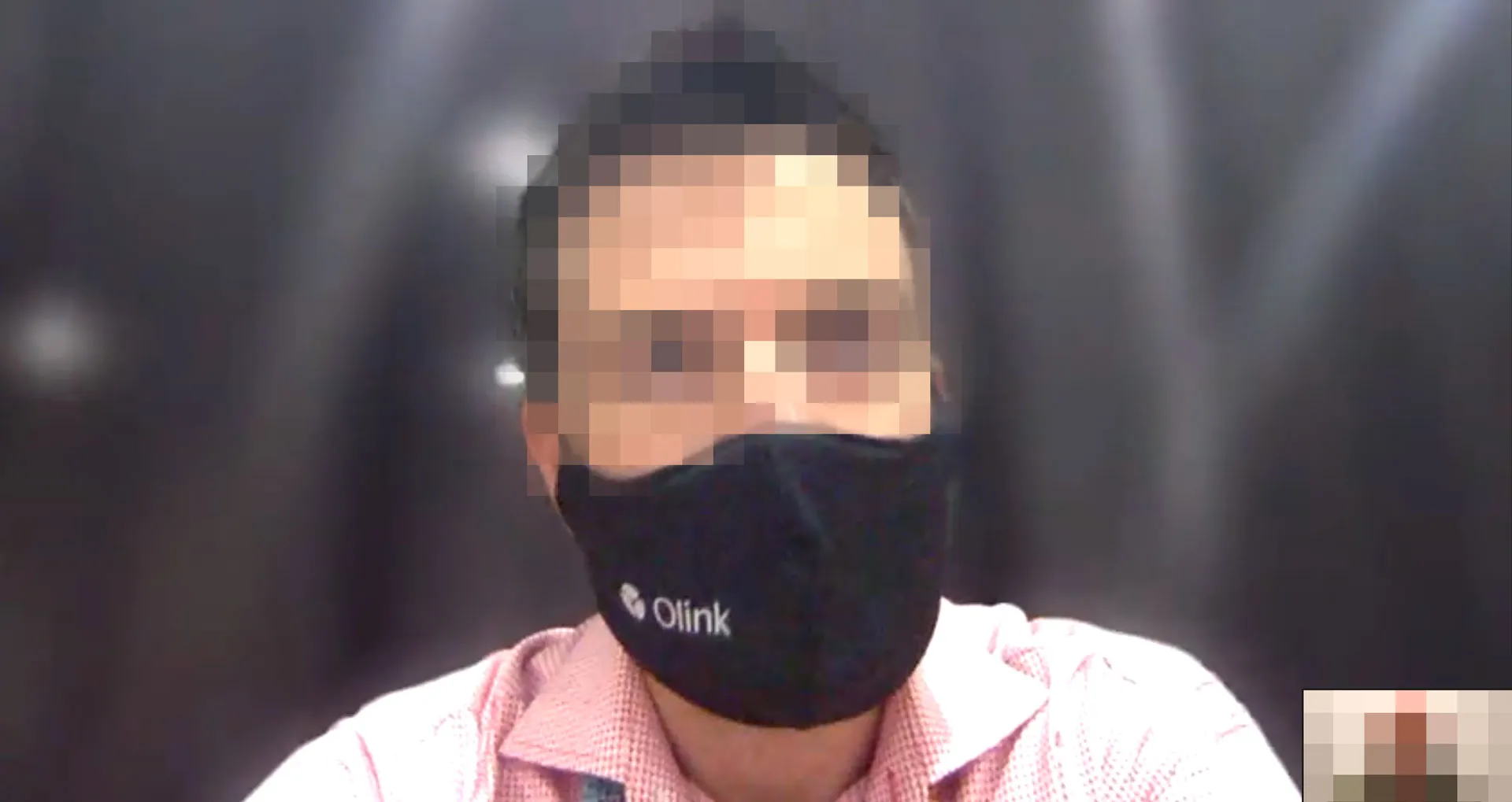 Video-call still of a person, face pixelated, wearing a black Olink-branded face mask