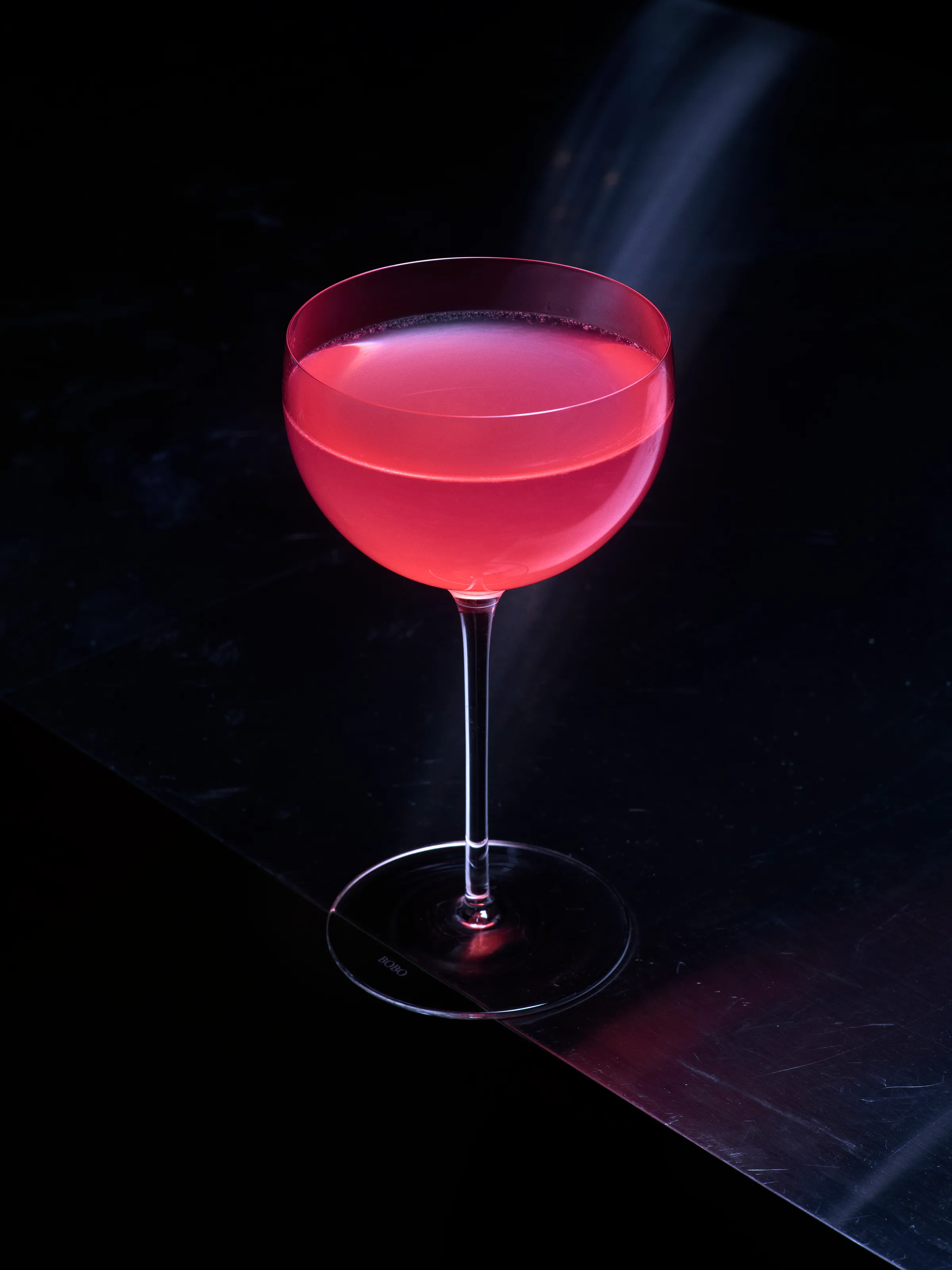 A pink cocktail in a stemmed coupe glass on a dark, scuffed metal bar top
