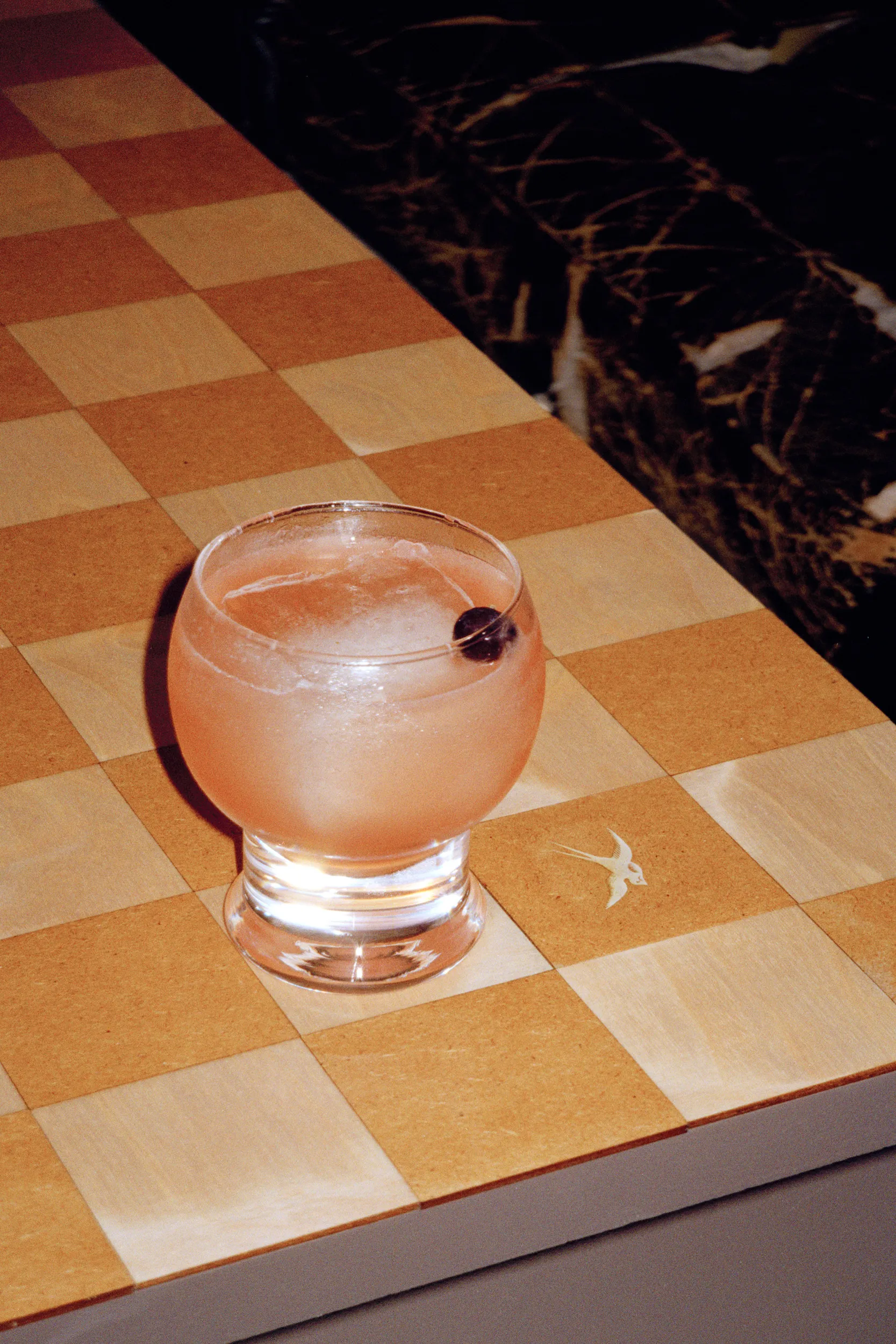A pink cocktail with a cherry on the checkered bar top, beside a swallow inlaid in one square
