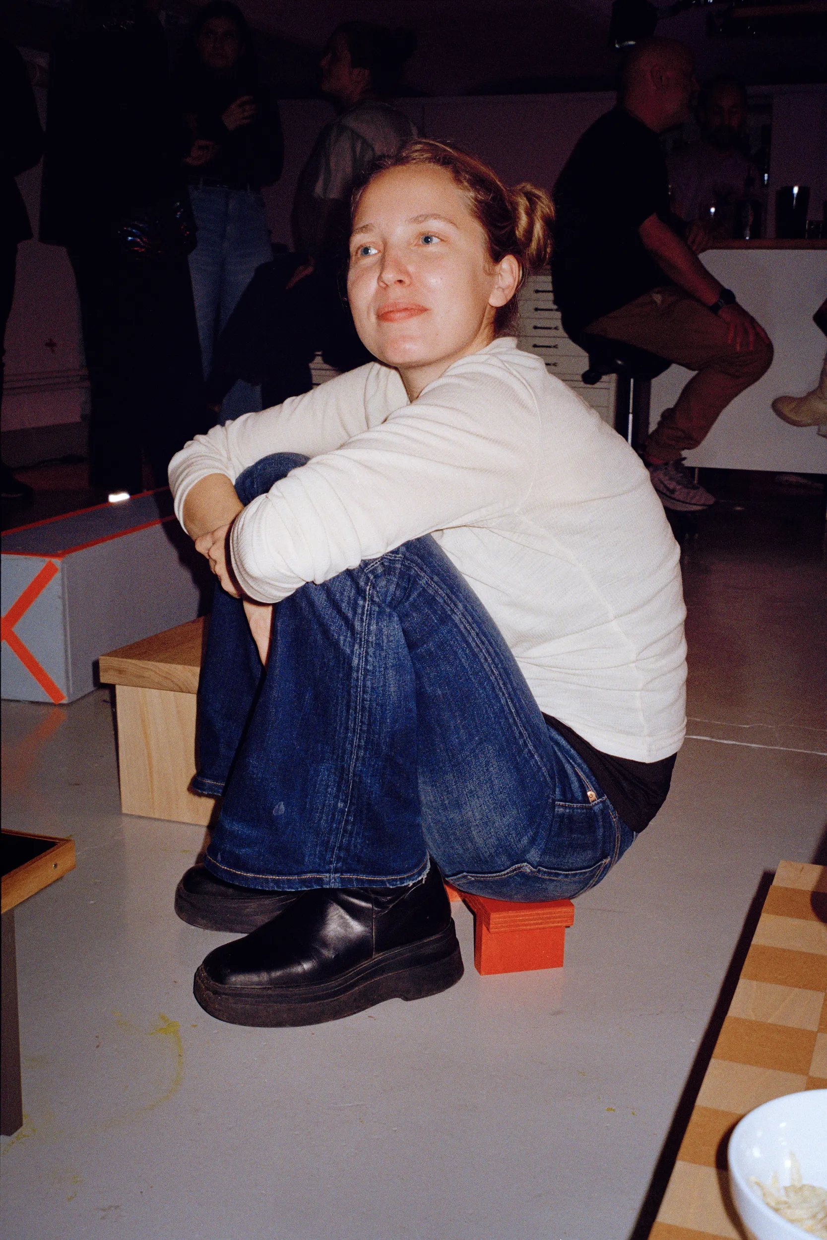 A guest in a white top and jeans sits hugging their knees on a small orange stool, other guests behind