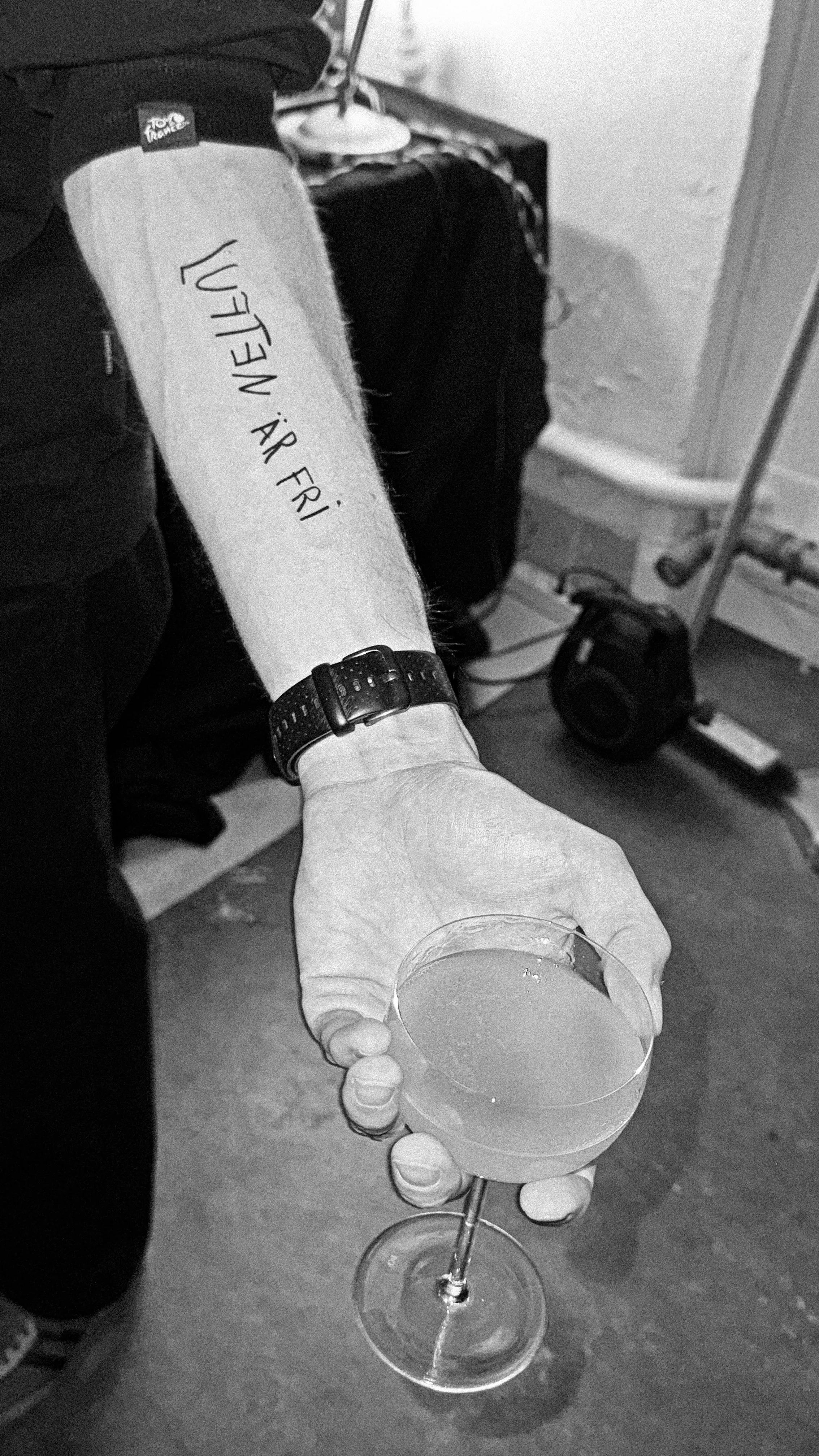 “Luften är fri” handwritten in black along a forearm, the hand holding a cocktail glass