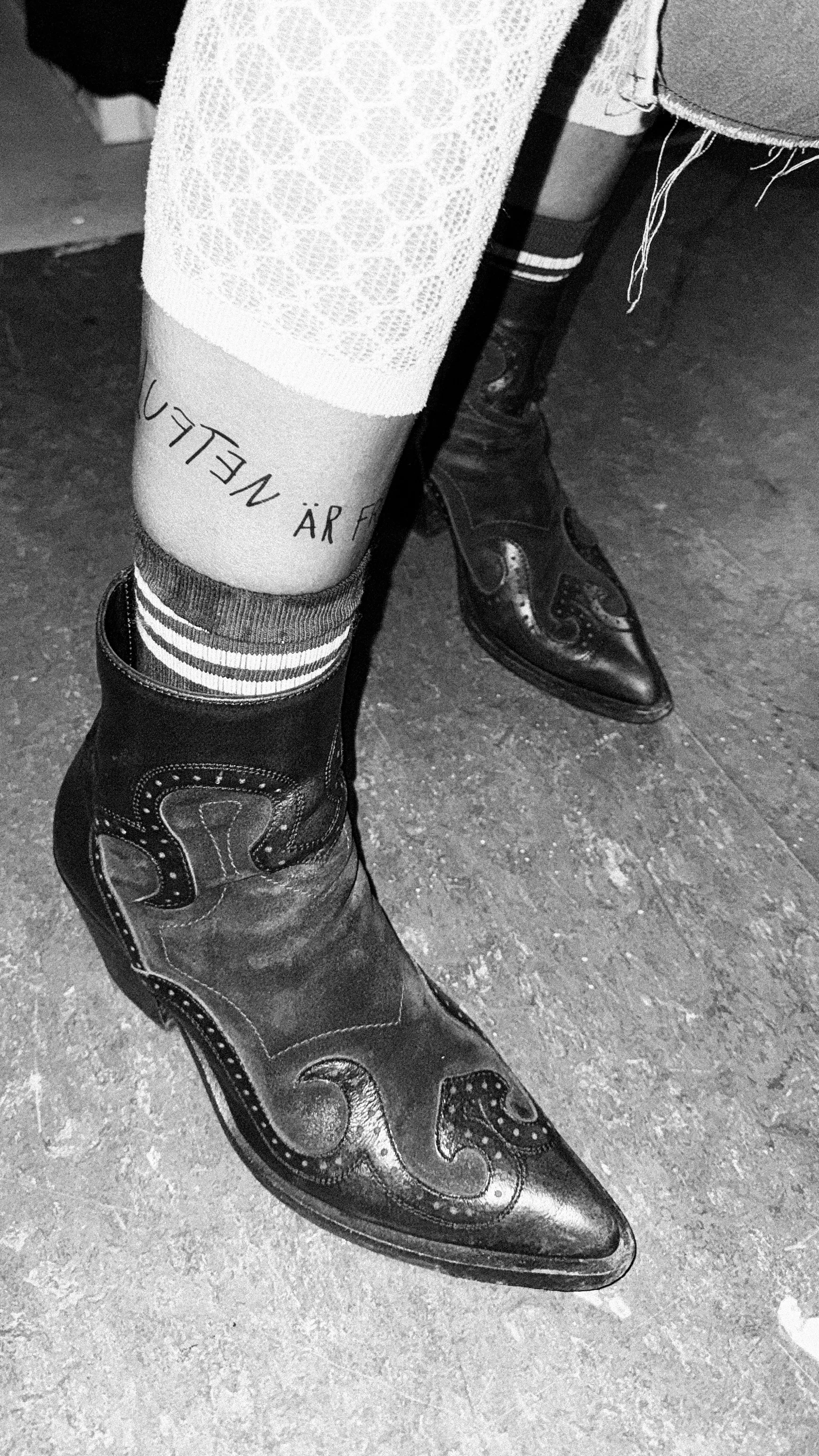 “Luften är fri” written around an ankle between a lace stocking and a striped sock, above a black cowboy boot