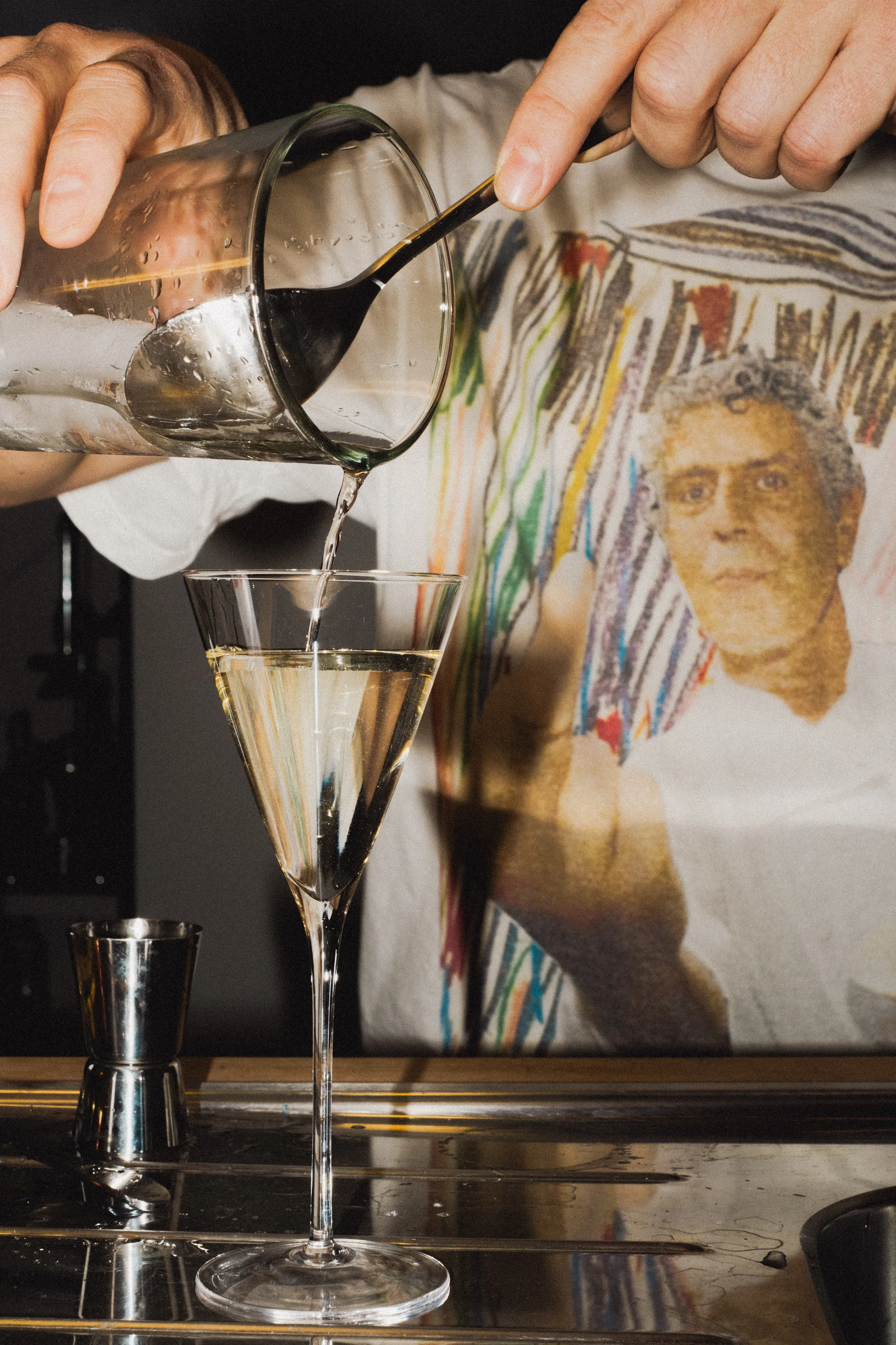 A bartender in a T-shirt printed with a painted portrait pours a drink from a mixing glass into a stemmed glass