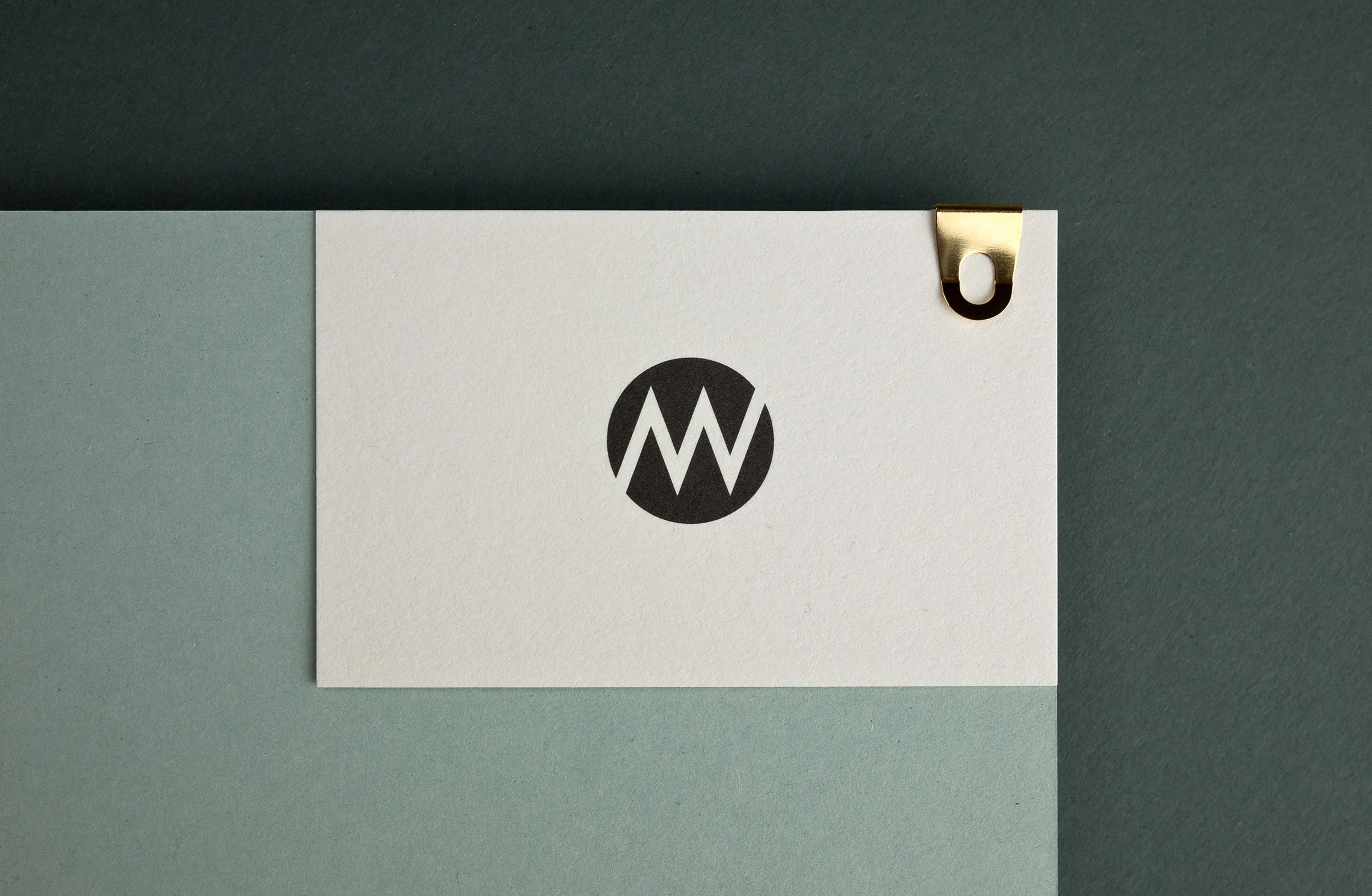 White card with a black circular M/W monogram and a gold clip, on sage and dark green paper